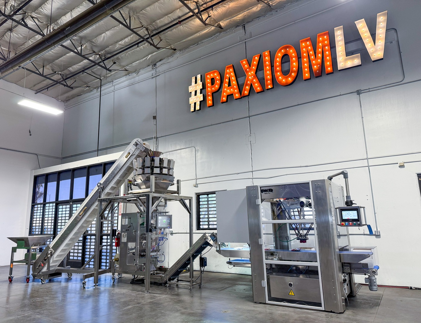 Complete bagging line at the Xperience Center: Primo Combi multihead weigher, vertical bagger, conveyance, and case packer beneath the #PAXIOMLV marquee